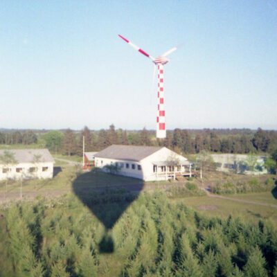 Tvindkraft wind turbine in the background with yellow buildings in front, photographed from a hot air ballon, the shadow of the ballon is visible in t he foreground